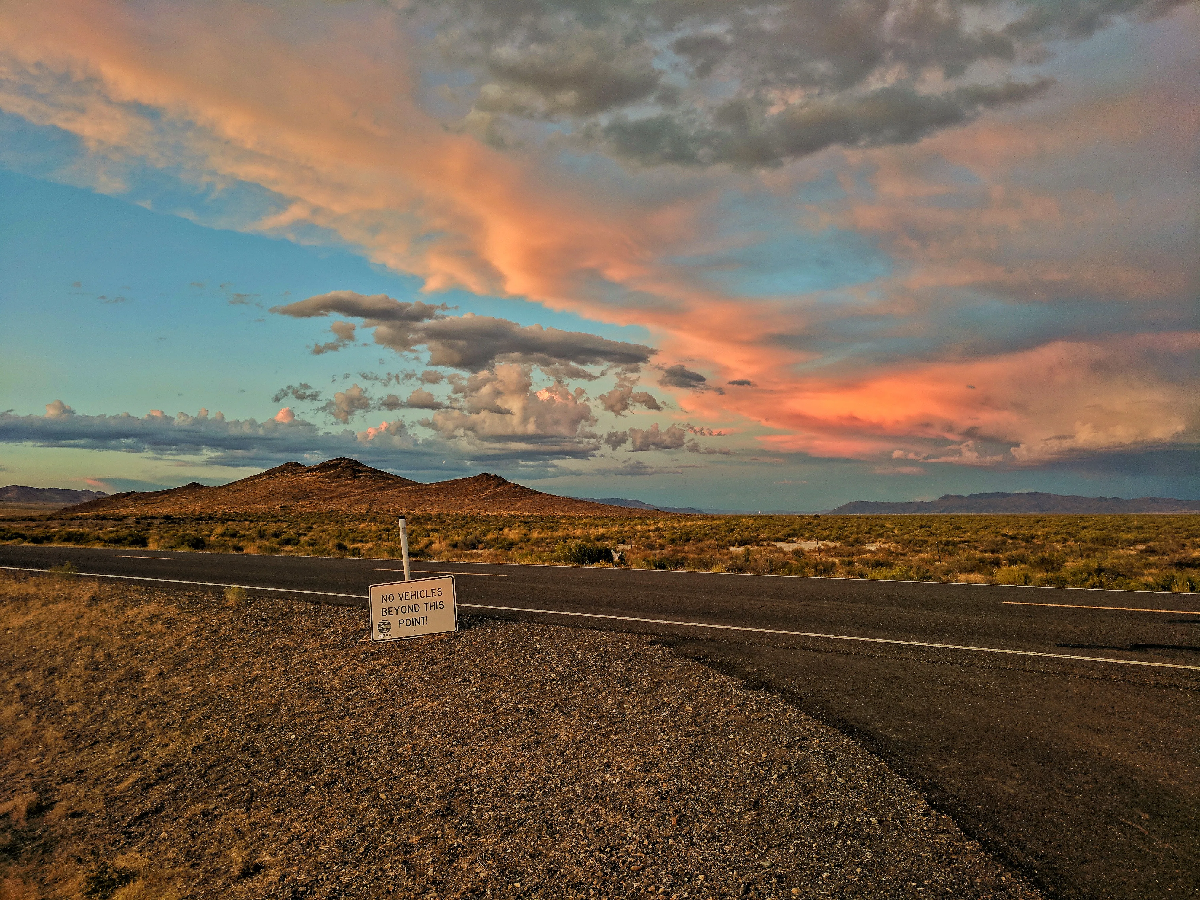 A "No Vehicles Beyond This Point" sign on the desert roadside, storm clouds lit pink and orange over distant mountains